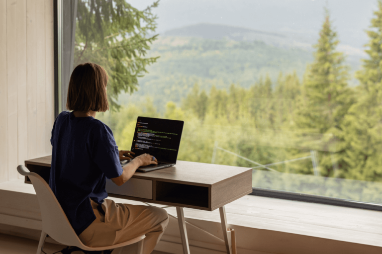 woman sitting at a desk with her laptop, facing a large window with a view of trees and hills