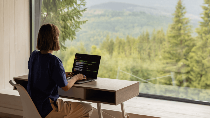 woman sitting at a desk with her laptop, facing a large window with a view of trees and hills