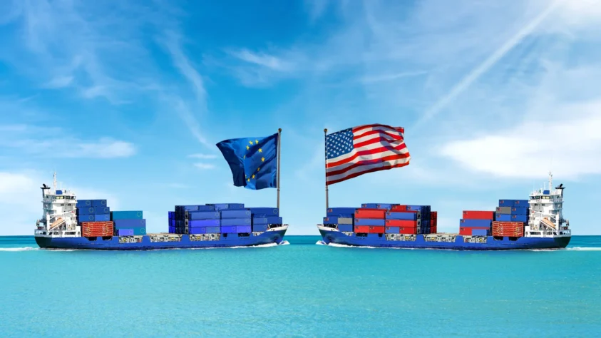 Two large cargo ships, one with an American flag and one with a European Union flag