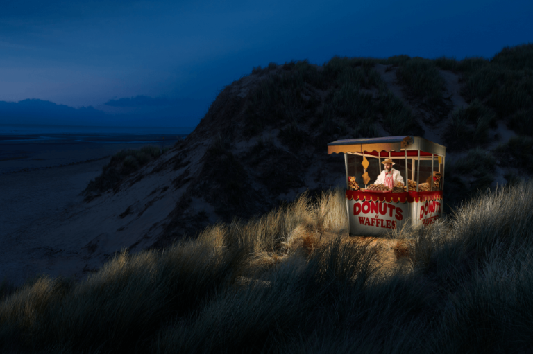 man in a carnival food stand, on the grassy dunes of a beach at nighttime