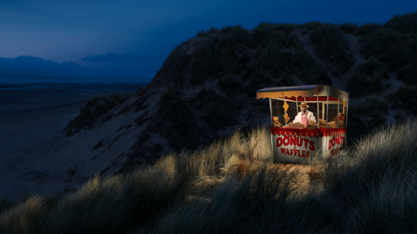 man in a carnival food stand, on the grassy dunes of a beach at nighttime